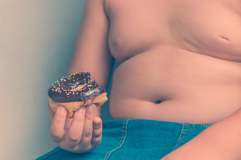 Diabesidade, uma combinação perigosa. Imagem de menino obeso, sem camiseta segurando um donut, representando o perigo da combinação entre diabetes e obesidade.