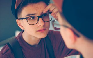 Puberdade Precoce - Imagem de menino criança olhando no espelho, de óculos, com muitas espinhas no rosto.