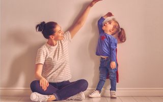 Mãe sentada no chão brincando de medir a filha, encostada na parede, vestida de super-heroina, para ilustrar o crescimento infantil.